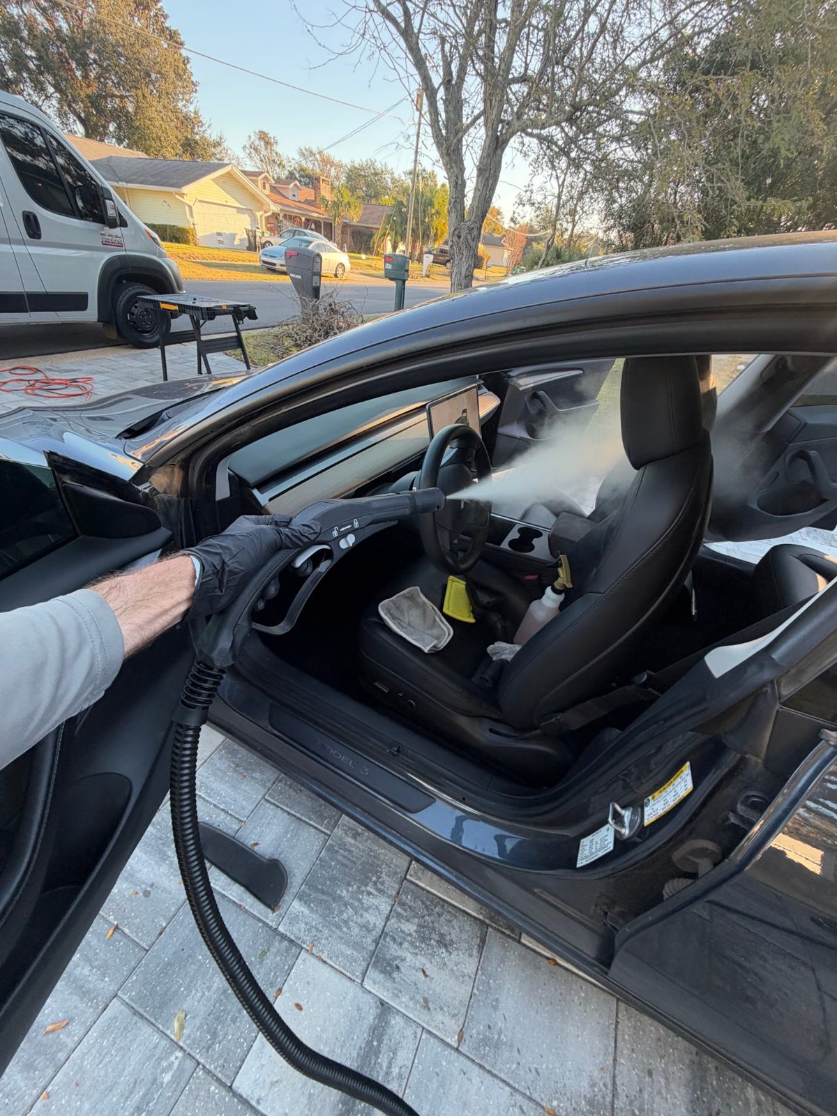 Gloved detailer steam cleaning the black leather driver's seat of a Tesla Model 3 with the door open in a driveway